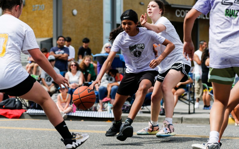A young girl playing basketball with another girl trying to block her and two others in the foreground. All are wearing gray team t-shirts.